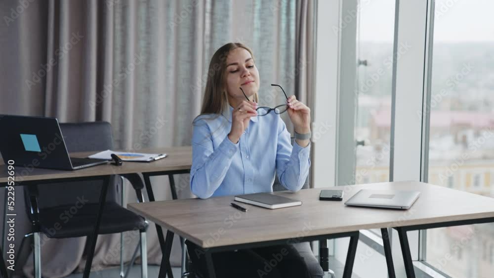 Thoughtful blonde lady sitting at desk puts on glasses. Woman looks straight ahead smiling happily. Office background. Thoughtful blonde lady sitting at desk puts on glasses. Woman looks straight ahead smiling happily. Office background.