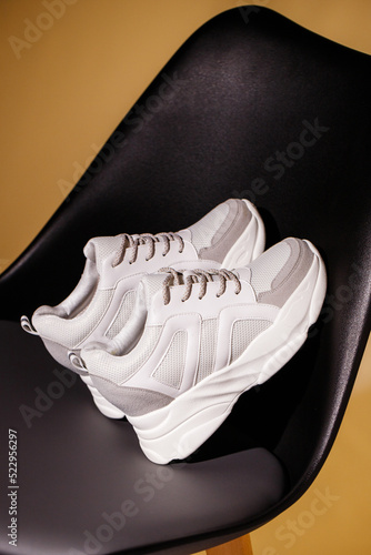 Close up of white sneakers leaning on back of black chair on beige studio background.