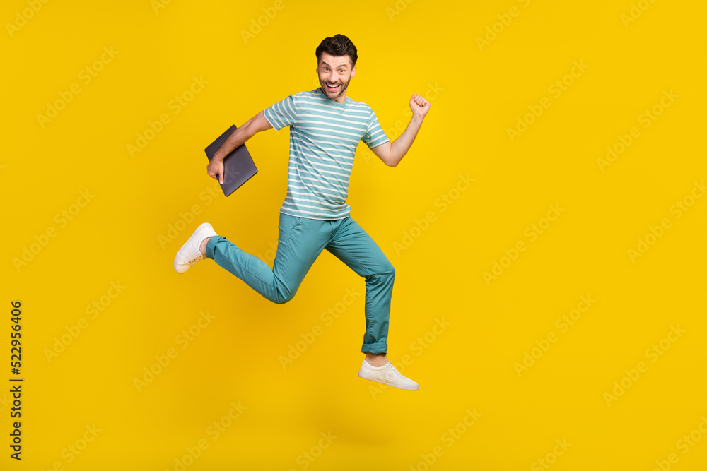 Full length photo of ecstatic handsome man wear striped t-shirt blue pants hold laptop run on vacation isolated on yellow color background
