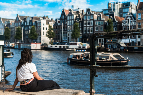 Mujer joven descansando sentada a orillas de un canal en un día soleado de verano en la ciudad de Ámsterdam. Un gran barco navegando por el canal frente a varios edificios típicos holandeses. © AliciaFdez