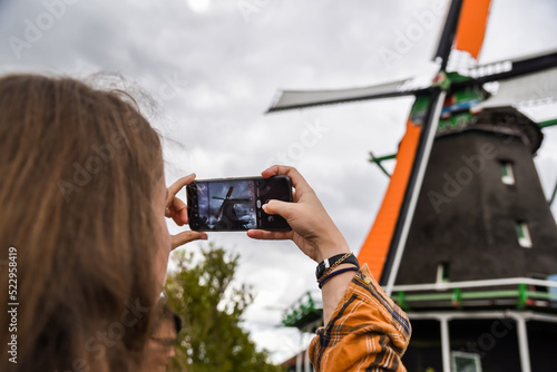 Una mujer rubia toma una fotografía con su teléfono móvil de un molino de viento en el pueblo de Zaanse Schans, Países Bajos. photo