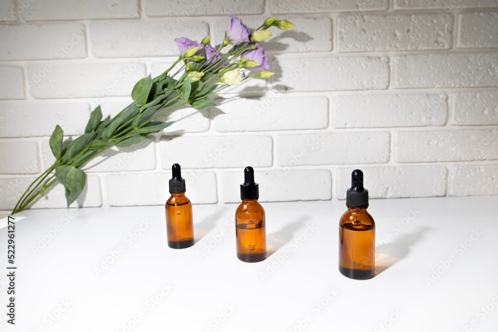 A group of amber bottle on a white table with face serum products near purple eustoma. Glass bottle with self care product. Front view A group of amber bottle on a white table with face serum products near purple eustoma. Glass bottle with self care product. Front view
