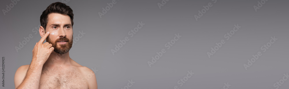 Obraz premium bearded and shirtless man applying cream on face isolated on grey, banner.