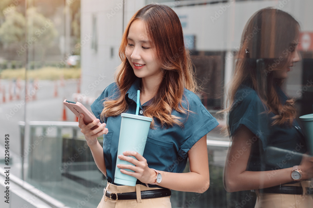 Smiling asian businesswoman use smartphone and hold reusable eco-friendly ecological cup while commuting in city in the morning Smiling asian businesswoman use smartphone and hold reusable eco-friendly ecological cup while commuting in city in the morning