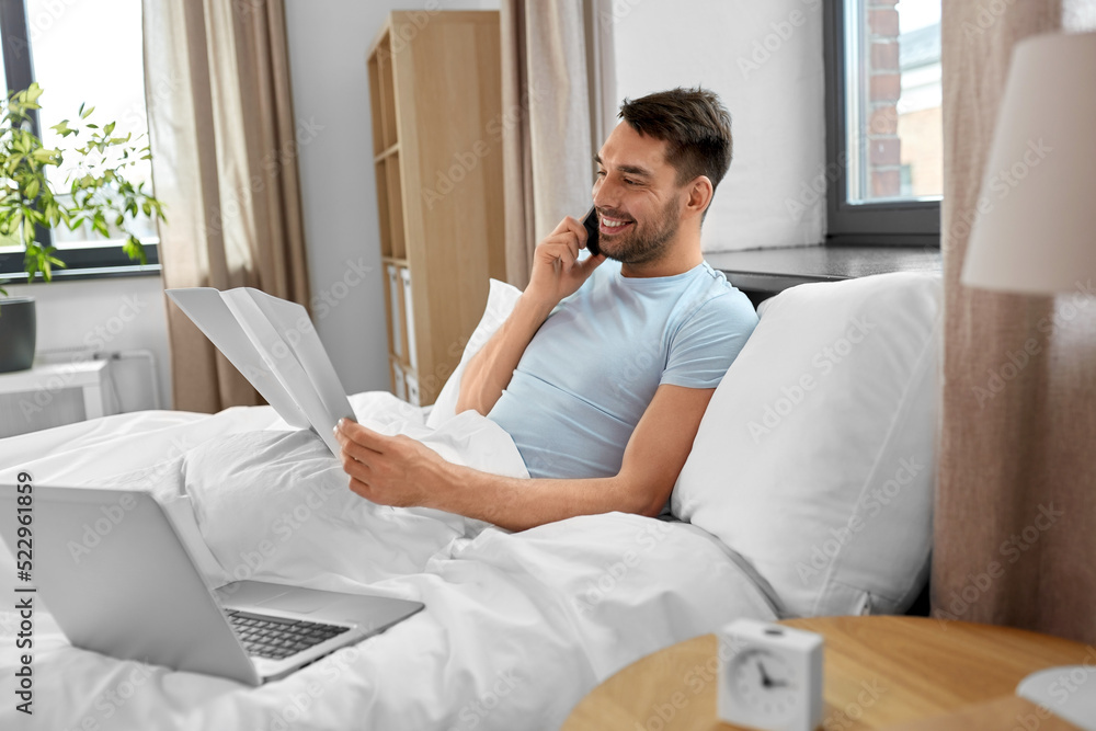 people, technology and remote job concept - man with folder and laptop computer calling on smartphone in bed at home bedroom people, technology and remote job concept - man with folder and laptop computer calling on smartphone in bed at home bedroom
