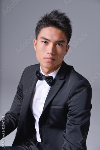 Handsome fashion model. elegant man wear formal black suit with bow tie sitting chair on gray background