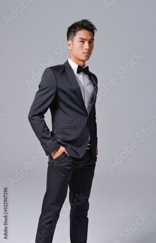 Handsome fashion model.
elegant man wear formal black suit with bow tie on gray background 

