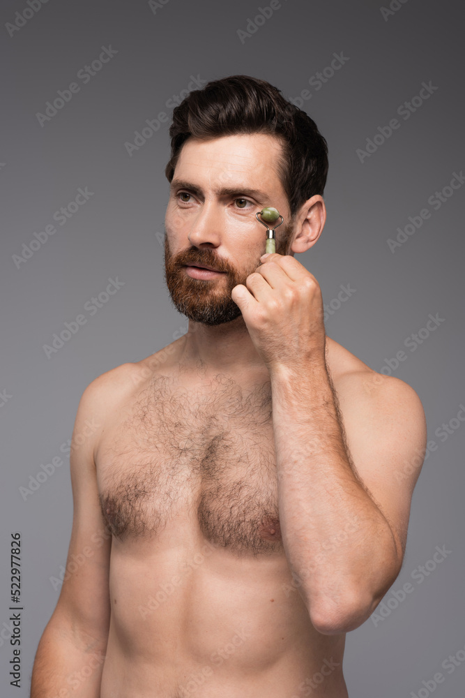 shirtless man with beard using jade roller while massaging face isolated on grey. shirtless man with beard using jade roller while massaging face isolated on grey.