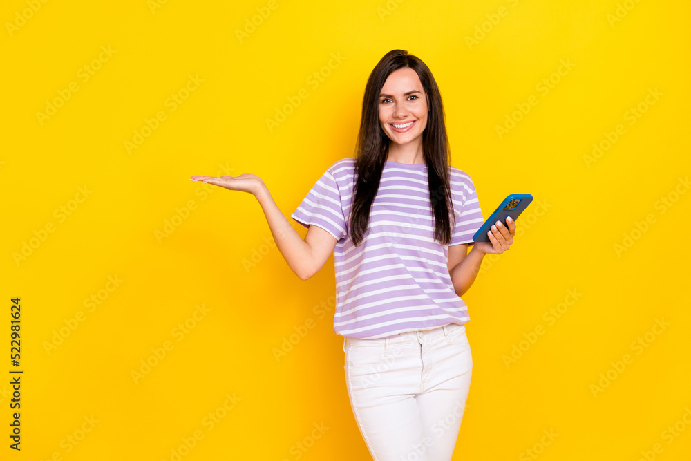 Photo of positive cute lady wear stylish striped outfit open arm hold telephone demonstrate empty space isolated on yellow color background Photo of positive cute lady wear stylish striped outfit open arm hold telephone demonstrate empty space isolated on yellow color background