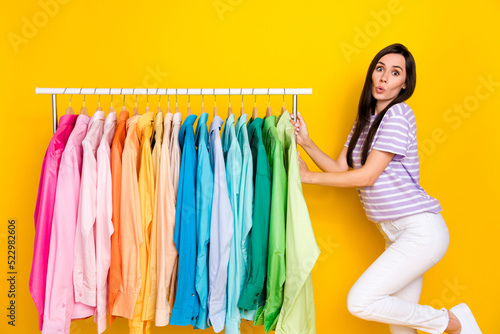 Portrait of pretty confused lady hand arm touch rack impressed reaction number trendy shirts isolated on yellow color background