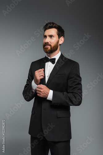 smiling man with beard standing in elegant suit with bow tie isolated on grey.