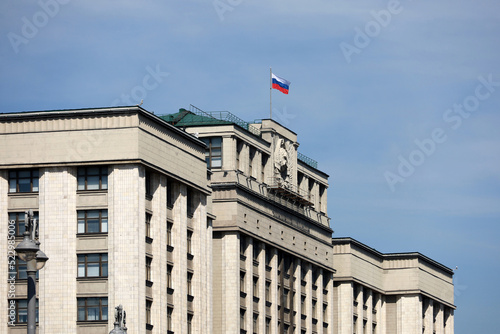 Parliament building in Moscow with Russian flag on background of blue sky. Facade of State Duma of Russia with soviet coat of arms, russian authority