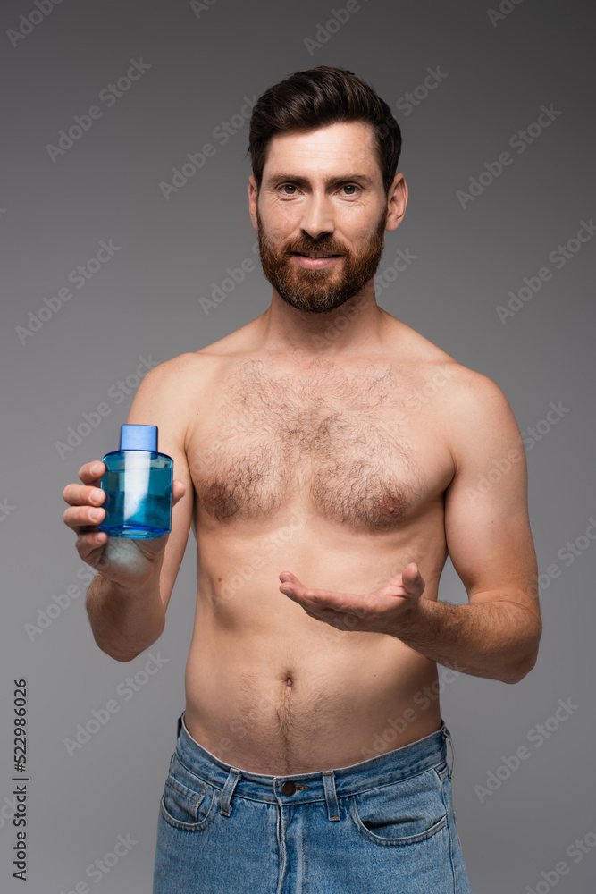 bearded and shirtless man pointing with hand at bottle with after shave product isolated on grey. bearded and shirtless man pointing with hand at bottle with after shave product isolated on grey.