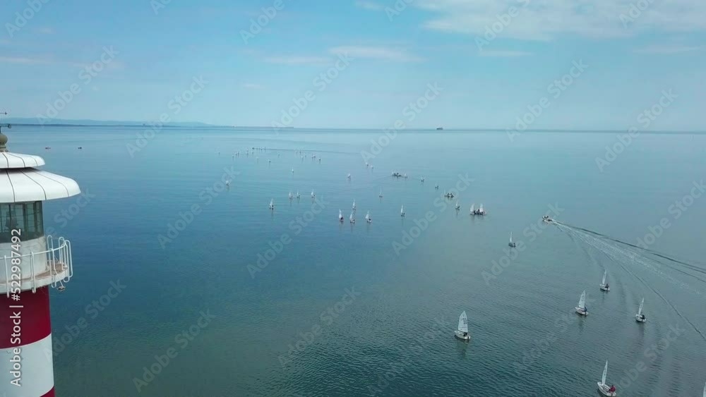 Towering Beacon Light Of Burgas With Sailing Small Sailboats In The Scenic Bulgarian Black Sea- International Regatta Participants. Aerial Lateral Shot Towering Beacon Light Of Burgas With Sailing Small Sailboats In The Scenic Bulgarian Black Sea- International Regatta Participants. Aerial Lateral Shot