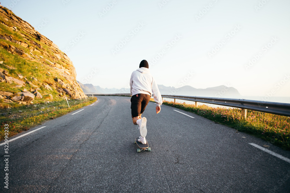 Hipster skater ride on longboard on empty epic road. Millennial man in hoodie and shorts ride skateboard in amazing location. Push to gain speed on longboard