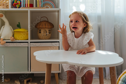 A little girl cries and is naughty sitting at the table in the nursery. Children's tantrums, tears and discontent. Problems of kid's upbringing. photo