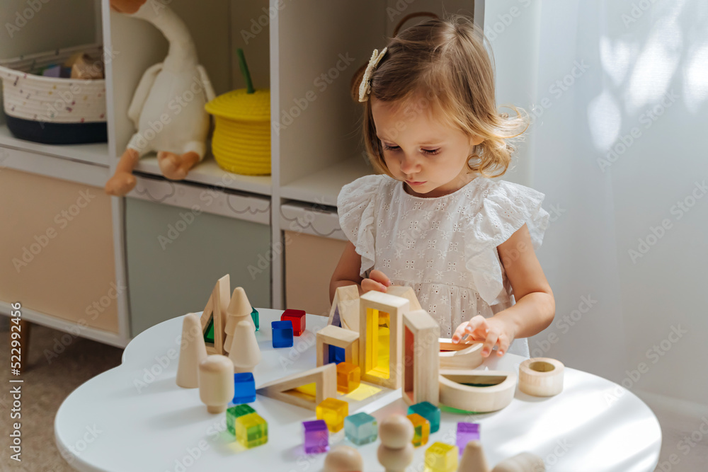 A little girl playing with wooden blocks on the table in playroom. Educational game for baby and toddler in modern nursery. The kid builds a tower from wooden rainbow stacking blocks. A little girl playing with wooden blocks on the table in playroom. Educational game for baby and toddler in modern nursery. The kid builds a tower from wooden rainbow stacking blocks.