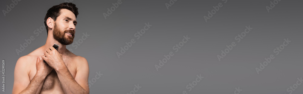 Obraz premium cheerful and shirtless man with hair on chest shaving with electric razor isolated on grey, banner.