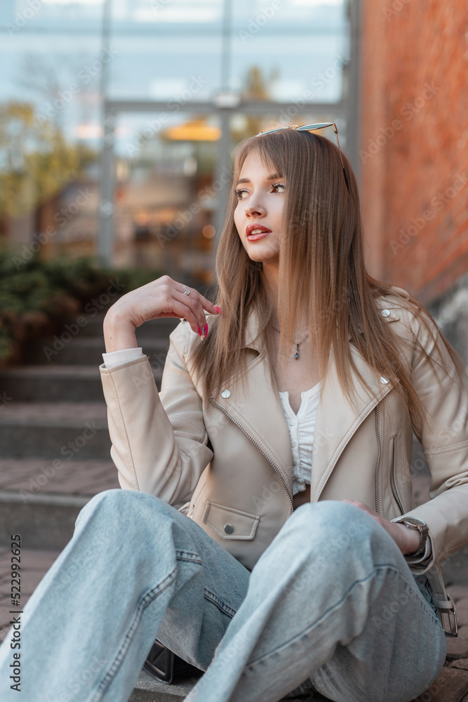 Trendy stylish beautiful young woman model in fashionable casual clothes with a leather jacket and vintage jeans sits on the steps near a modern red brick building mall Trendy stylish beautiful young woman model in fashionable casual clothes with a leather jacket and vintage jeans sits on the steps near a modern red brick building mall