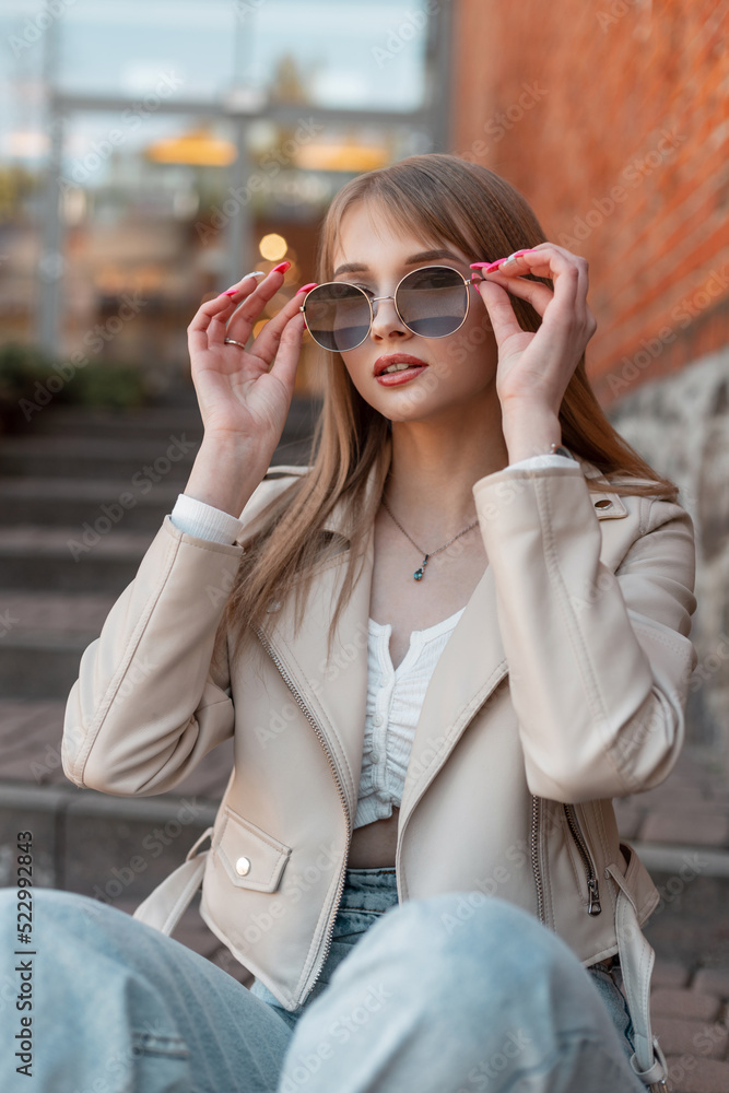 Cool pretty young zoomer woman with leather fashion white jacket and jeans wears a stylish sunglasses sits on the steps and rest near a vintage brick mall building Cool pretty young zoomer woman with leather fashion white jacket and jeans wears a stylish sunglasses sits on the steps and rest near a vintage brick mall building