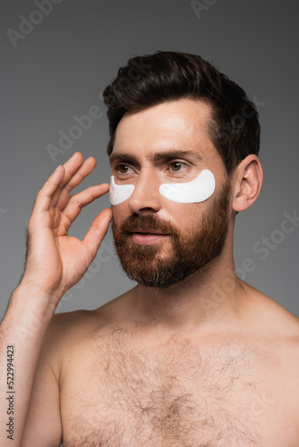 bearded man applying moisturizing eye patches isolated on grey.