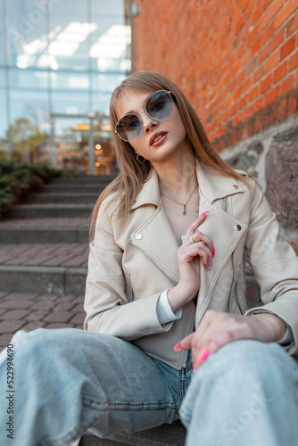 Street fashionable young pretty woman with stylish casual autumn outfit with fashion sunglasses, leather jacket, top and jeans sits near a brick building mall © alones
