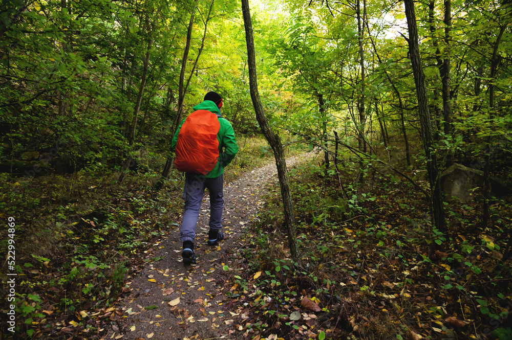 A tourist in a green jacket and a red backpack walks along a mountain path in spring or autumn. Traveler, tourist in autumn landscape. Mountains and trekking, rear view A tourist in a green jacket and a red backpack walks along a mountain path in spring or autumn. Traveler, tourist in autumn landscape. Mountains and trekking, rear view