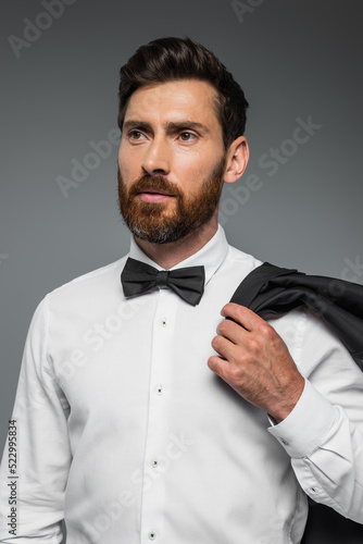 portrait of bearded man in suit with bow tie holding blazer isolated on grey.