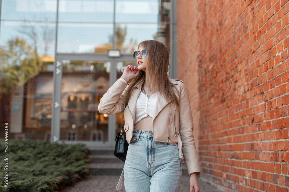 Beautiful woman with leather jacket, fashion jeans, top and stylish bag walks in the city and puts on a vintage sunglasses. Trendy urban female style look clothes