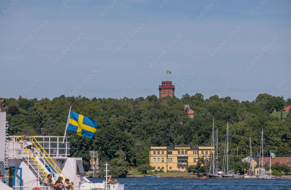 Stern of a commuting boat with Swedish flag passing the island Djurgården, a brick tower with flag and sailboats in a harbor, a sunny summer day in Stockholm