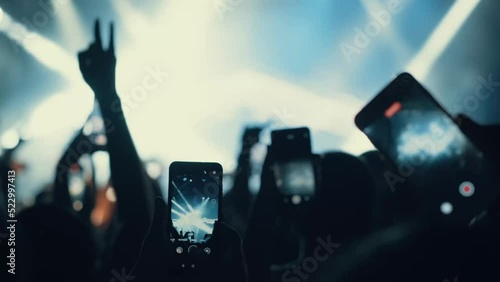 A blurry and illuminated dancing crowd. A crowd of fans standing near the stage applauding their favorite rock band. Concert in the city center. Young people with smartphones. Silhouettes of musicians