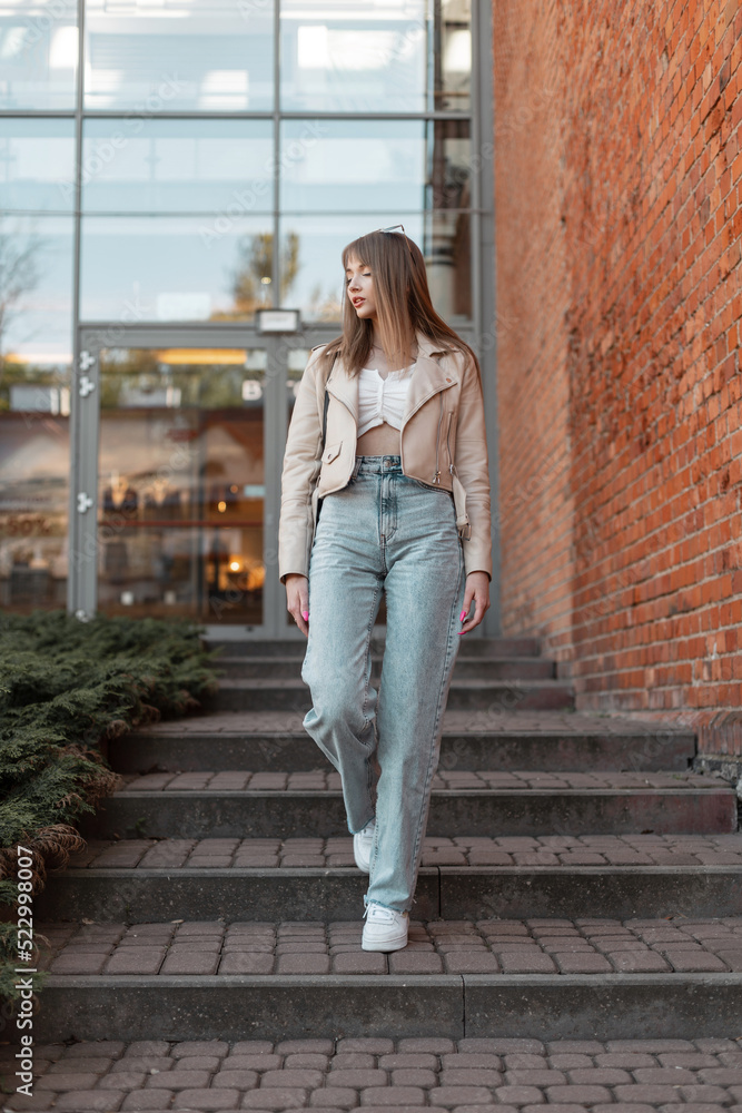 Trendy beautiful young woman in fashionable casual wear outfit with a leather jacket with bell-bottom high-waisted jeans and white sneakers walks near a red brick store building Trendy beautiful young woman in fashionable casual wear outfit with a leather jacket with bell-bottom high-waisted jeans and white sneakers walks near a red brick store building