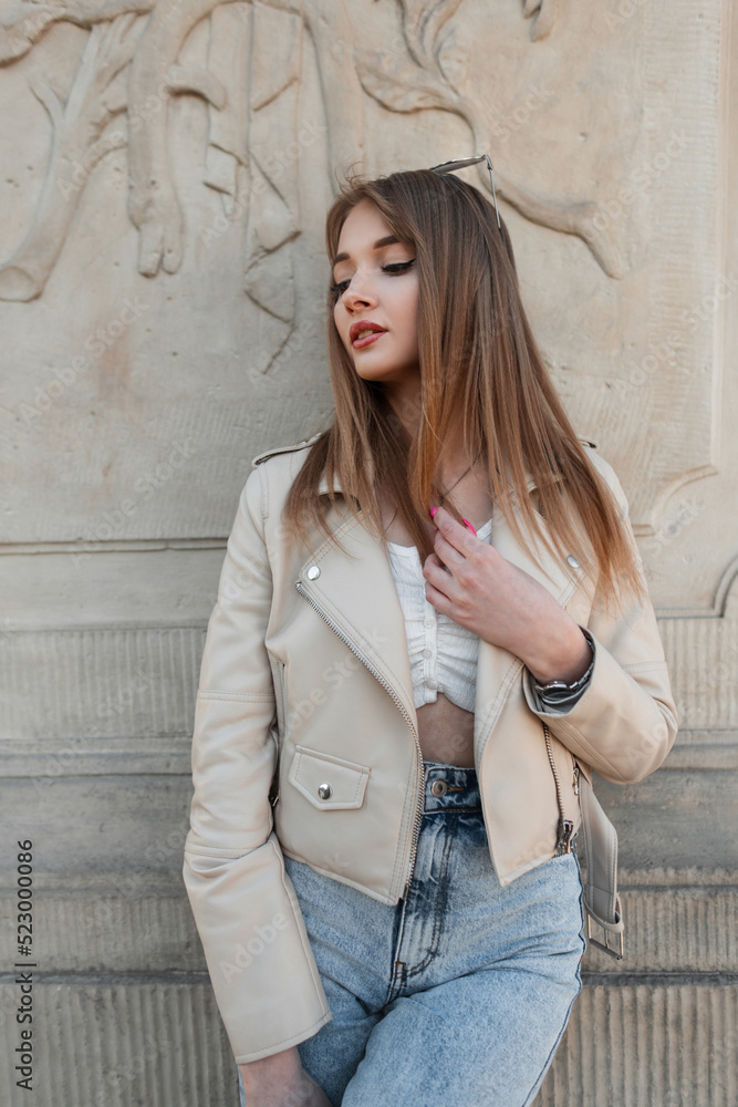 Stylish young woman model in a fashionable leather jacket with a top and high-waisted jeans stands near a vintage column. Women's street style, fashion and beauty