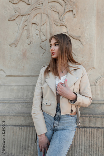 Pretty young beauty woman hipster with hair in fashionable leather jacket and top with vintage blue jeans stands near a wall on the street © alones