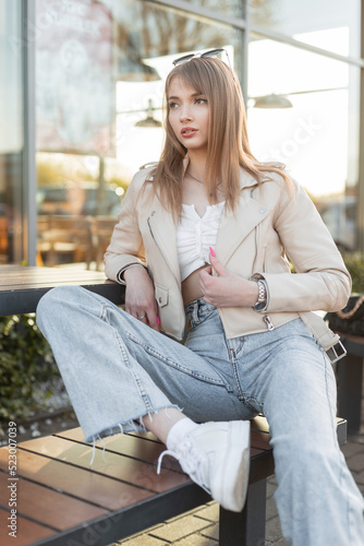 Stylish pretty young Caucasian woman in urban streetwear with leather rock jacket, top, blue jeans and white sneakers sits on wooden bench near a cafe in the city at sunset