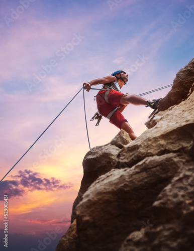 Tablou canvas Muscular climber man in protective helmet abseiling from cliff rock wall using rope Belay device and climbing harness on evening sunset sky background