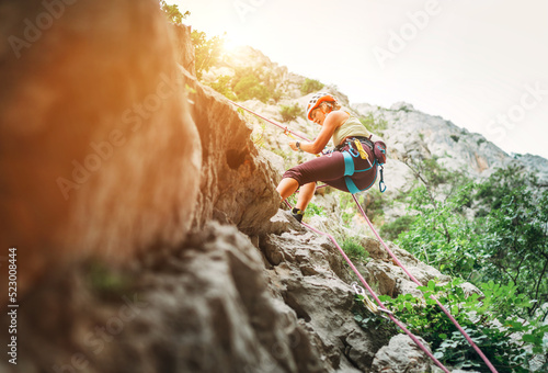 Slika na platnu Active climber woman in protective helmet abseiling from cliff rock wall using rope with belay device and climbing harness