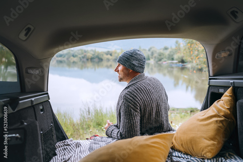 Midle-aged smiling man dressed in warm knitted clothes sitting in the opened Car trunk and enjoying a beautiful autumnal mountain lake view after night. Warm early autumn auto traveling concept image.