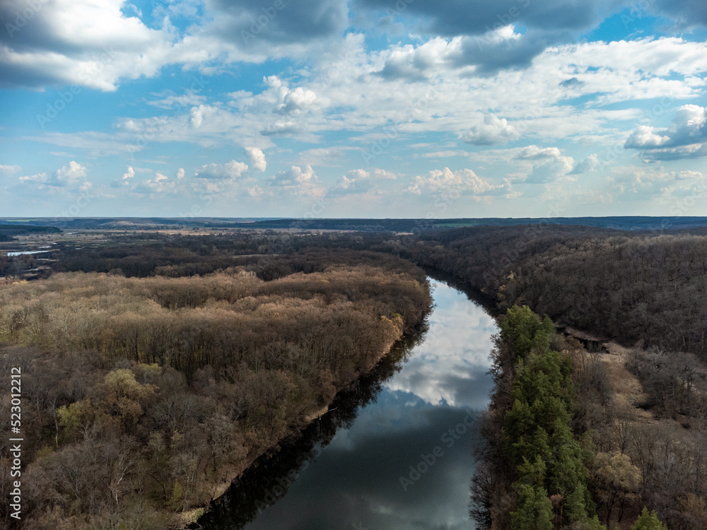 Aerial view on river curve in bare trees forest. Epic blue cloudscape reflection in water. Sunny nature landscape from drone