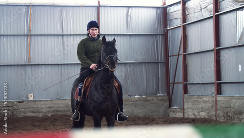 in special hangar, a young disabled man learns to ride a black, thoroughbred horse, hippotherapy. man has an artificial limb instead of his right leg. concept of rehabilitation of disabled with
