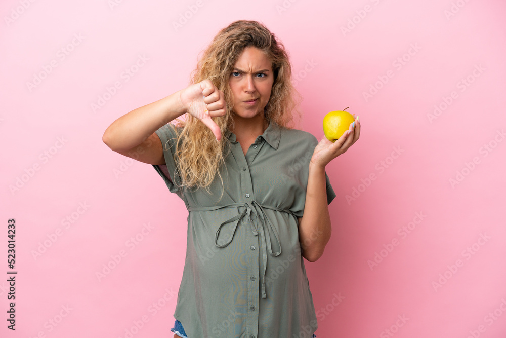 Girl with curly hair isolated on pink background pregnant and holding an apple while doing bad signal Girl with curly hair isolated on pink background pregnant and holding an apple while doing bad signal