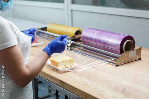 Woman wraps piece of cheese lying on plastic tray in transparent food film.