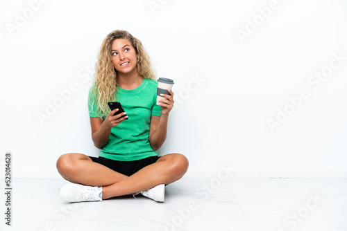 Girl with curly hair sitting on the floor holding coffee to take away and a mobile while thinking something