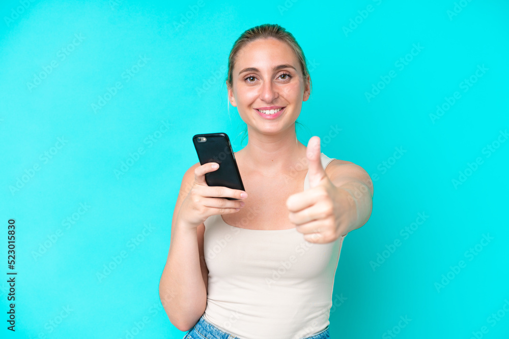 Young caucasian woman isolated on blue background using mobile phone while doing thumbs up Young caucasian woman isolated on blue background using mobile phone while doing thumbs up