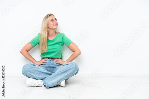 Young caucasian woman sitting on the floor isolated on white background posing with arms at hip and smiling