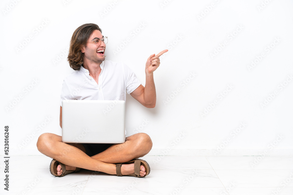 Young handsome man with a laptop sitting on the floor isolated on white background pointing finger to the side and presenting a product Young handsome man with a laptop sitting on the floor isolated on white background pointing finger to the side and presenting a product