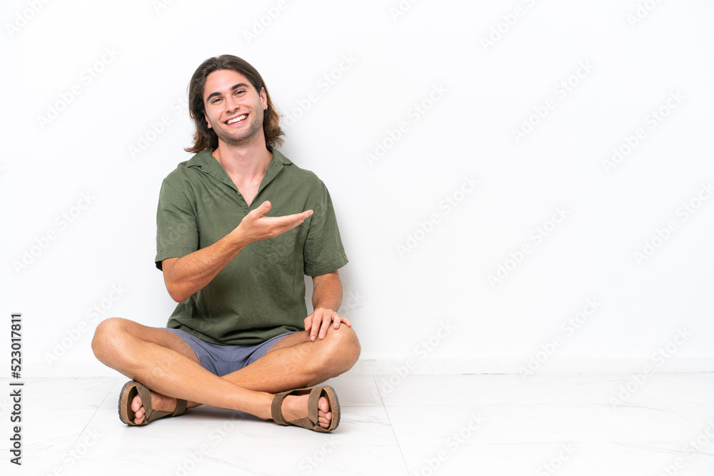 Young handsome man sitting on the floor isolated on white background presenting an idea while looking smiling towards Young handsome man sitting on the floor isolated on white background presenting an idea while looking smiling towards