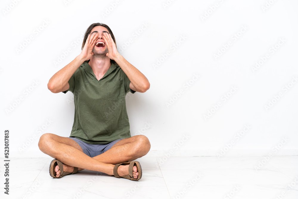 Young handsome man sitting on the floor isolated on white background shouting and announcing something Young handsome man sitting on the floor isolated on white background shouting and announcing something