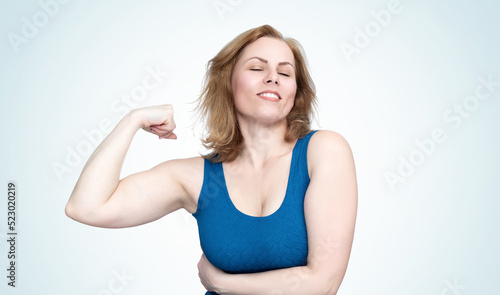 Young woman in a tight blue dress shows the muscles on her arm, smiling broadly and closing her eyes, isolated on light blue background