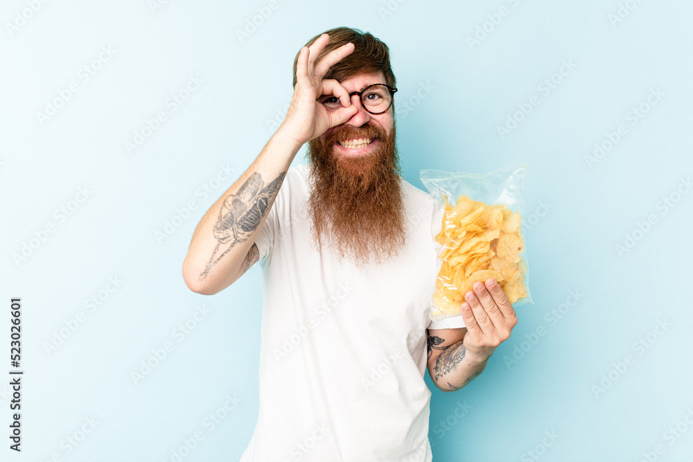 Young caucasian man holding a bag of chips isolated on blue background excited keeping ok gesture on eye. Young caucasian man holding a bag of chips isolated on blue background excited keeping ok gesture on eye.